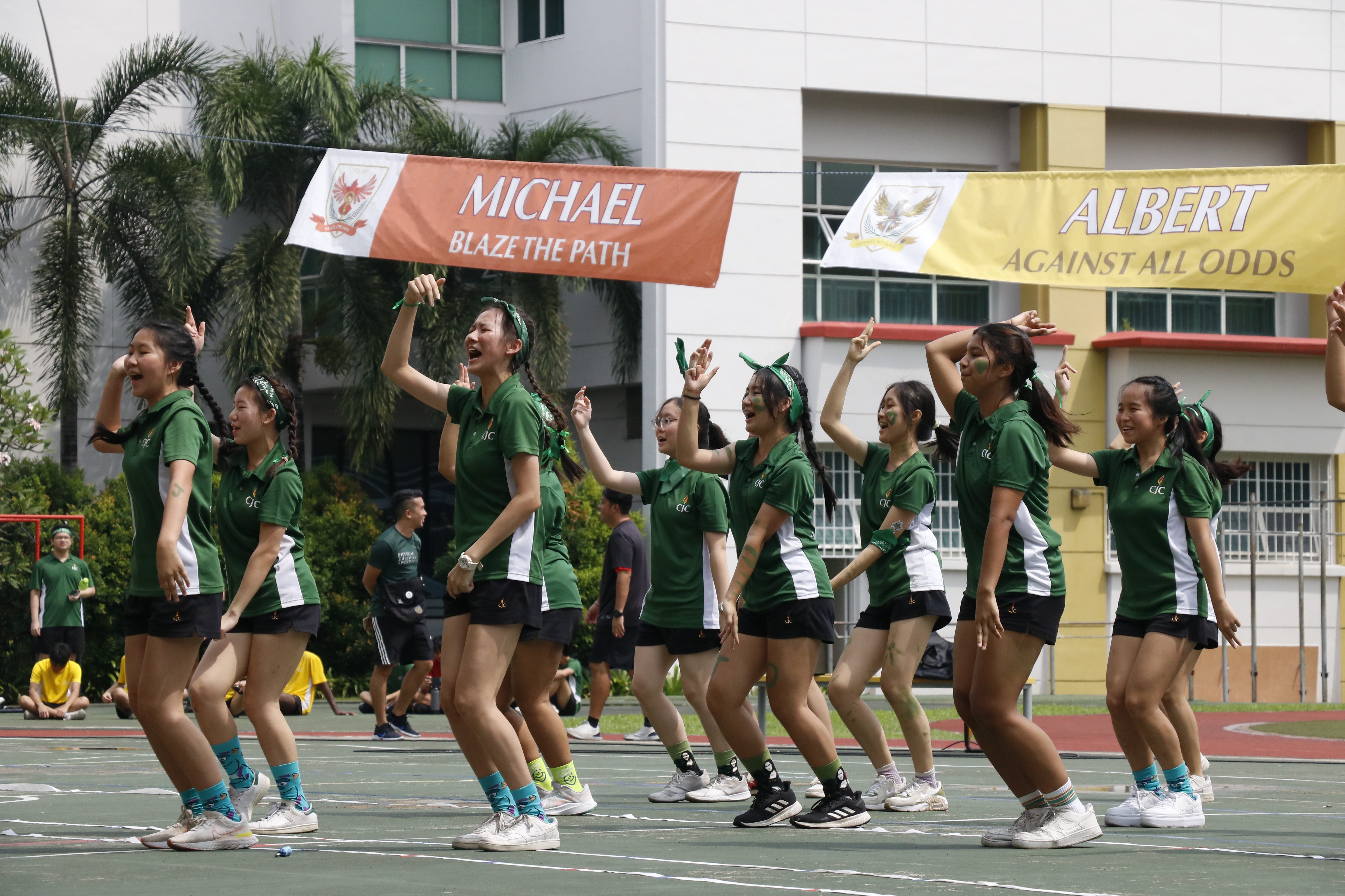 Group of women in green jerseys cheer and dance on a court beneath banners reading MICHAEL and ALBERT.